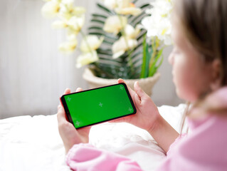 A teenage girl lies on the bed and holds a phone with a green screen in her hands in her beautiful...