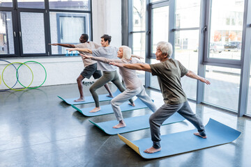 Senior asian man in warrior pose practicing yoga near multiethnic friends in sports center.