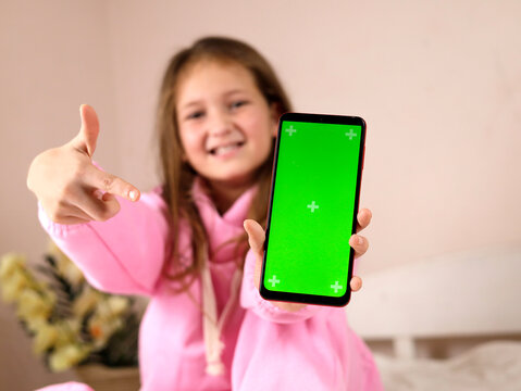 A beautiful and smiling teenage girl in a pink suit sits on her bed at home and holds a phone with a green screen with finger pointing at the screen. used for advertising, business, goods. Soft focus