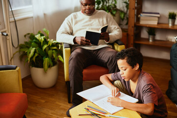 Smart African boy doing his math homework in the living room while his grandpa in the blurry...