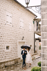 Fototapeta na wymiar Bride and groom walk past the stone buildings on the cobblestones under an umbrella