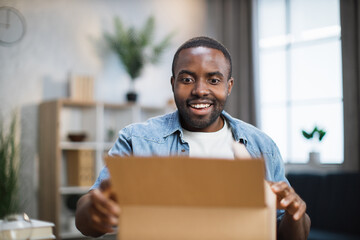 Excited male influencer opening box with ordered goods while staying at home. African american man...