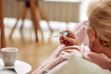 Close up of sick elderly woman checking her temperature with thermometer lying on sofa caughing...