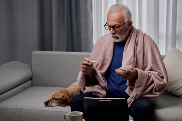 Sick elderly man checking his temperature suffering from seasonal flu or cold and using tablet for...