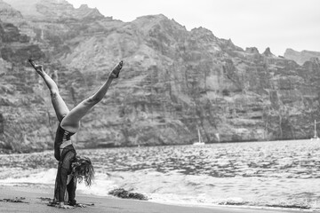 Young fitness woman doing handstand exercise on the rocky beach, practicing yoga on seashore....