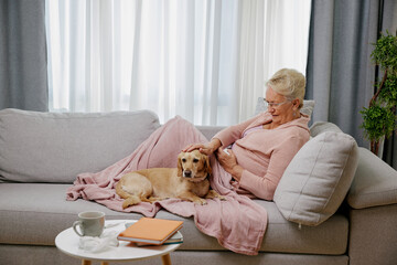 Sick elderly woman suffering from seasonal flu or cold lying down on sofa with her small dog. Ill senior feel unhealthy with influenza at home