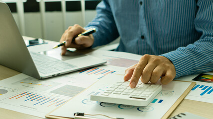 An accountant presses a calculating calculator to make a monthly financial report or company profit at his desk with graph papers and a work-from-home ideal laptop.