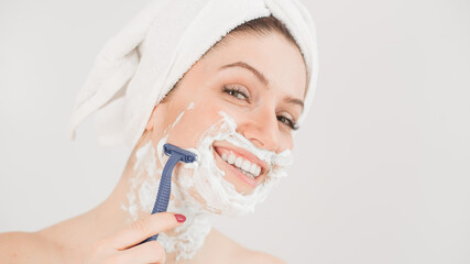 Cheerful caucasian woman with a towel on her head and shaving foam on her face holds a razor on a...
