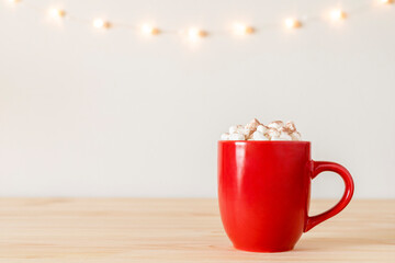 Hot chocolate in red mug with small marshmallows on wooden table with holiday lights on wall. Cocoa in a cup, hot winter drink