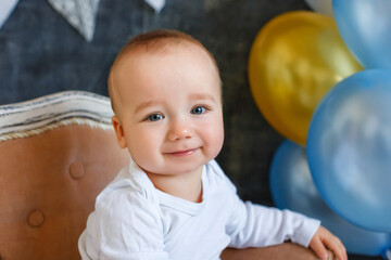 Portrait of a little boy in the studio. The child is sitting on a mini-chair near a bunch of balloons on a dark gray background decorated with a garland of flags.