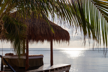 chaise à massage dans une cabane avec un toit de paille sur un bord de mer avec le soleil qui reflète dans l'eau à l'horizon
