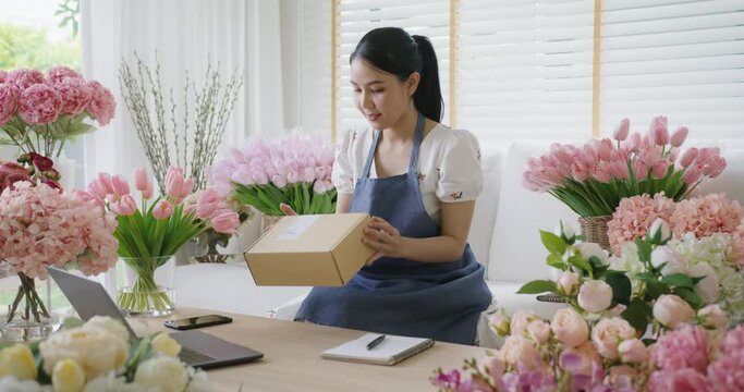 Asia people SME owner young woman smile enjoy selling prepare sale order product wrap flower gift box carton send parcel by post service. Happy freelance job work at home in digital modern store shop.