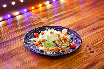 Caesar salad in a dark clay plate on a wooden table with multicolored lights on the background.