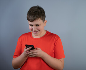 A smiling young man in a red T-shirt looks at his smartphone, reads text messages. A teenager with a phone, isolated on a gray background