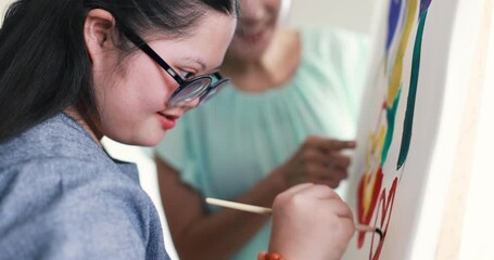 Delighted child with down syndrome creating painting on canvas and celebrating goal achievement with raised hands during art lesson with Asian teacher while looking at camera