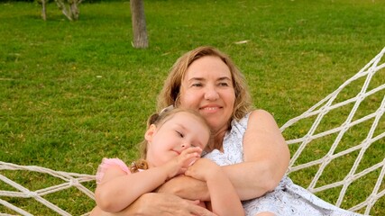 cute couple grandmother and granddaughter 3 years old are relaxing and basking in a hammock on a green meadow in their garden on a summer sunny day. Vacation concept, generational relationship