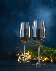 Two transparent champagne glasses on a dark blue textured table. Fir branches with garlands in the background. Festive atmosphere