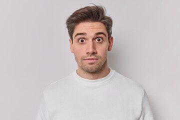 Fototapeta premium Portrait of shocked startled adult man stares bugged eyes at camera hears big news dressed in casual jumper isolated over white background. Concerned young guy feels very surprised poses indoor