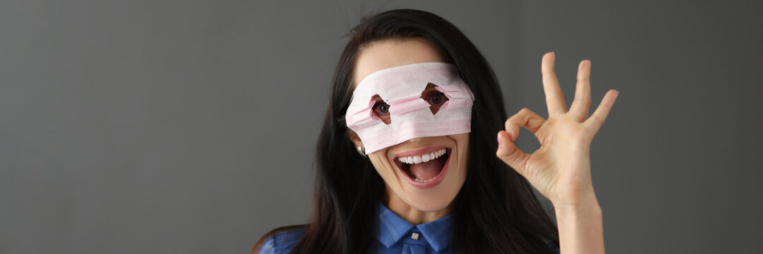 Young woman looking through holes on medical protective mask and showing hand ok gesture