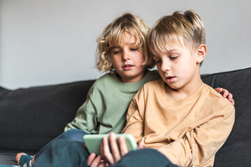 Low angle of calm little sibling with blond hair in casual outfits watching interesting video on smartphone. while sitting together on comfortable sofa at home
