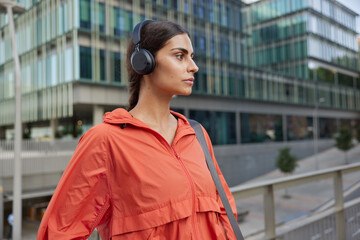 Horizontal shot of thoughtful sporty brunette European woman wears wireless headphones listens music being on her way to gym dressed in anorak poses against blurred background in urban setting © WHstudio Leushin N