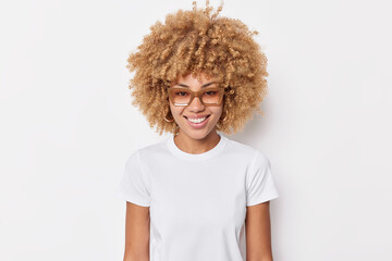Portrait of pretty young woman with curly hair smiles happily wears spectacles casual basic t shirt...