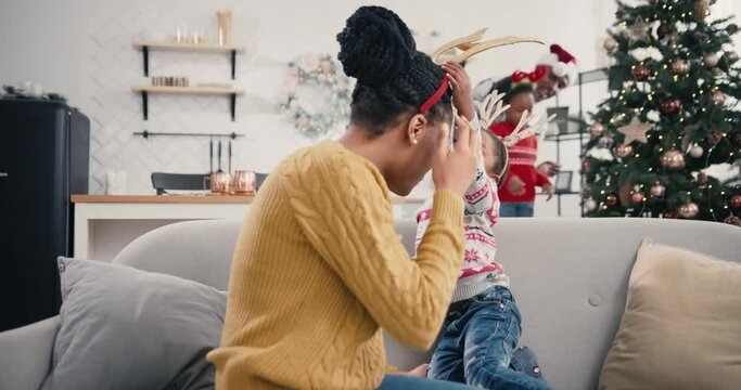 Portrait of mother playing with little cute child while sitting on sofa in christmassy decorated house. Happy handsome loving African American mom and kid spending time together. Christmas concept