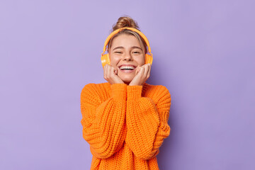 Portrait of cheerful young pretty woman smiles happily keeps hands under chin smiles broadly enjoys spare time for listening music wears casual orange jumper uses wireless headphones poses indoor