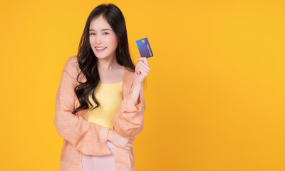 Cheerful asian woman wearing casual clothes while showing credit card mockup in own hand while standing with copy space over isolated yellow background. Payment purchase and financial concept.