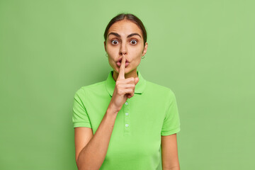 Impressed brunette woman makes silence gesture presses index finger over lips stunned to hear...