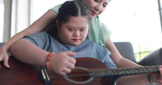 Loving Asian woman teaching child with down syndrome playing acoustic guitar while spending time together at home