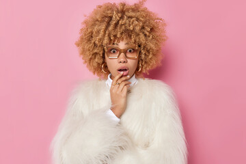 Shocked embarrassed young woman with curly hair keeps mouth opened cannot believe in surprising news wears spectacles and white fur coat isolated over pink background. Human reactions concept