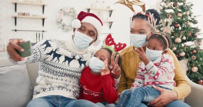 Portrait of happy African American family taking pictures on smartphone and smiling in christmassy decorated home. Dad in Santa hat taking selfie photo on cellphone with kids and wife in face masks