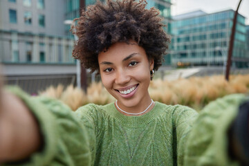 Young pretty woman takes selfie outdoors strolls in street smiles happily wears casual green jumper...