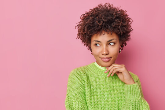 Horizontal shot of thoughtful young woman with curly hair keeps hand under chin concentrated away pensively ponders on something wears knitted sweater isolated over pink background blank space