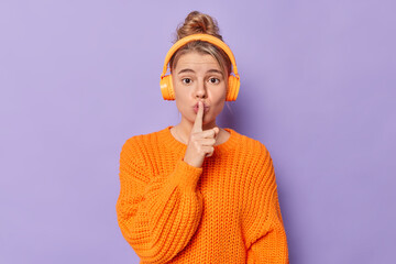 Mysterious young lovely woman makes silence gesture keeps index finger over lips tells secret listens audio track via wireless headphones wears knitted sweater isolated over purple background