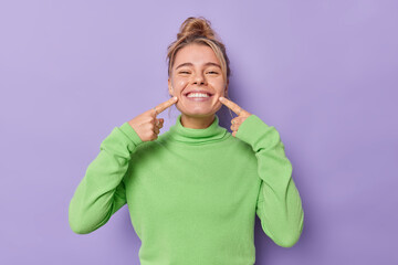 Beautiful happy fair haired European woman points index fingers at mouth forces cheerful smile shows perfect white teeth wears green jumper being in good mood isolated over purple background.