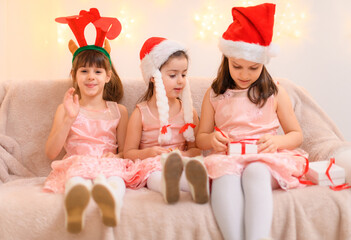 portrait of girls celebrating christmas or new year, children dressed in santa helper hat, sitting on a couch in home interior decorated with christmas lights and new year holiday gifts