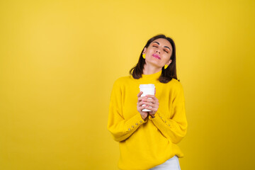 Young woman in a yellow cozy sweater with bright pink lip gloss with a paper cup of hot aromatic coffee, dreamily looks, cute smile, radiating warmth