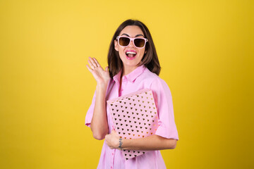 Stylish young woman in a pink dress and glasses on a yellow background with a gift box in her hands celebrates the best holiday, birthday, Valentine's Day