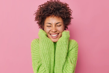 Portrait of good looking young woman smiles tenderly dressed in long sleeved jumper closes eyes recalls nice memory expresses joy isolated over pink background. Happy emotions and feelings concept