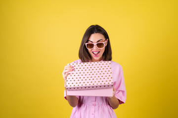 Stylish young woman in a pink dress and glasses on a yellow background with a gift box in her hands celebrates the best holiday, birthday, Valentine's Day