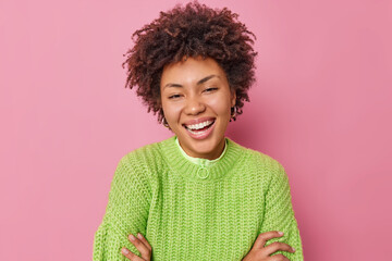 Portrait of happy curly haired young woman smiles positively wears casual green knitted sweater has upbeat mood expresses positive emotions poses against pink background hears pleasant news.