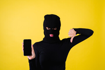 Woman with bright pink lips, balaclava on her head, holding a mobile phone, hacker, internet fraudster, pointing her finger at a blank black screen, showing thumbs down dissatisfied