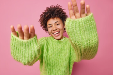 Playful happy woman keeps hands forwards camera smiles joyfully wears warm knitted jumper expresses...