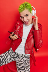 Naklejka premium Vertical shot of young good looking woman with trendy hairstyle listens music via stereo headphones holds mobile phone sits on comfortable chair isolated over red background. Technology concept