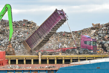 Machine operated grabbing arm, loading scrap metal onto a ship in the harbour of Newhaven, UK, with lorry delivering more scrap metal to the dock yard.
