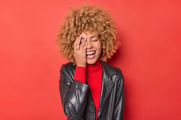 Positive young woman with fair curly hair makes face palm feels overjoyed laughs happily wears turtleneck and black leather jacket isolated over vivid red background. Sincere authentic emotions