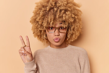 Lovely cheeky beautiful young woman with curly bushy hair makes peace sign keeps lips folded wears casual jumper poses indoor against beige background for cute photograph. Body language concept