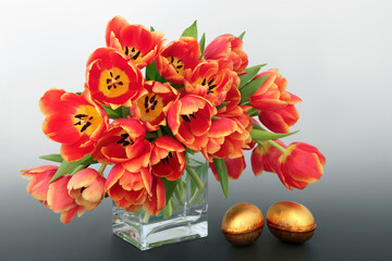 Gold Easter eggs with red and yellow spring tulip flowers on gradient grey background. Springtime and Easter still life beautiful nature concept.